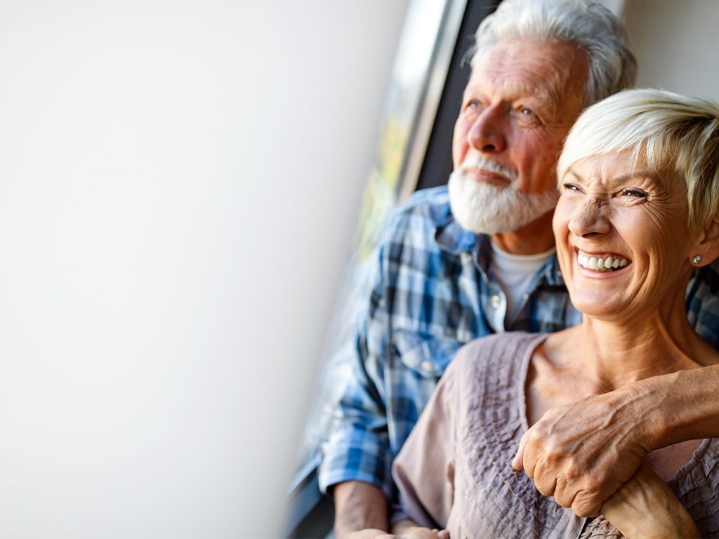 Image of two older people holding hands, smiling, and looking out the window. 